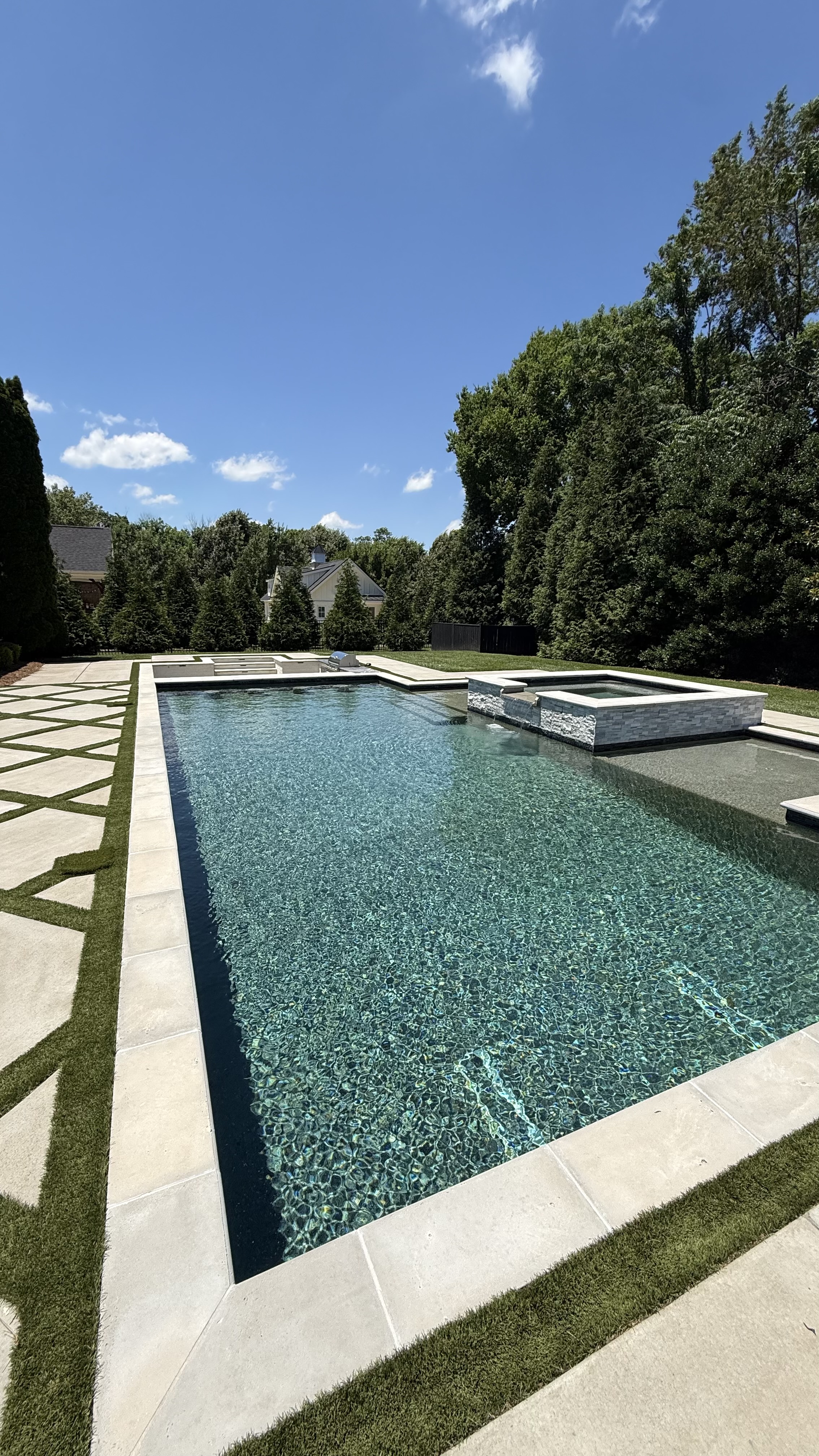 Bright outdoor swimming pool with a stone patio, lawn, and trees
