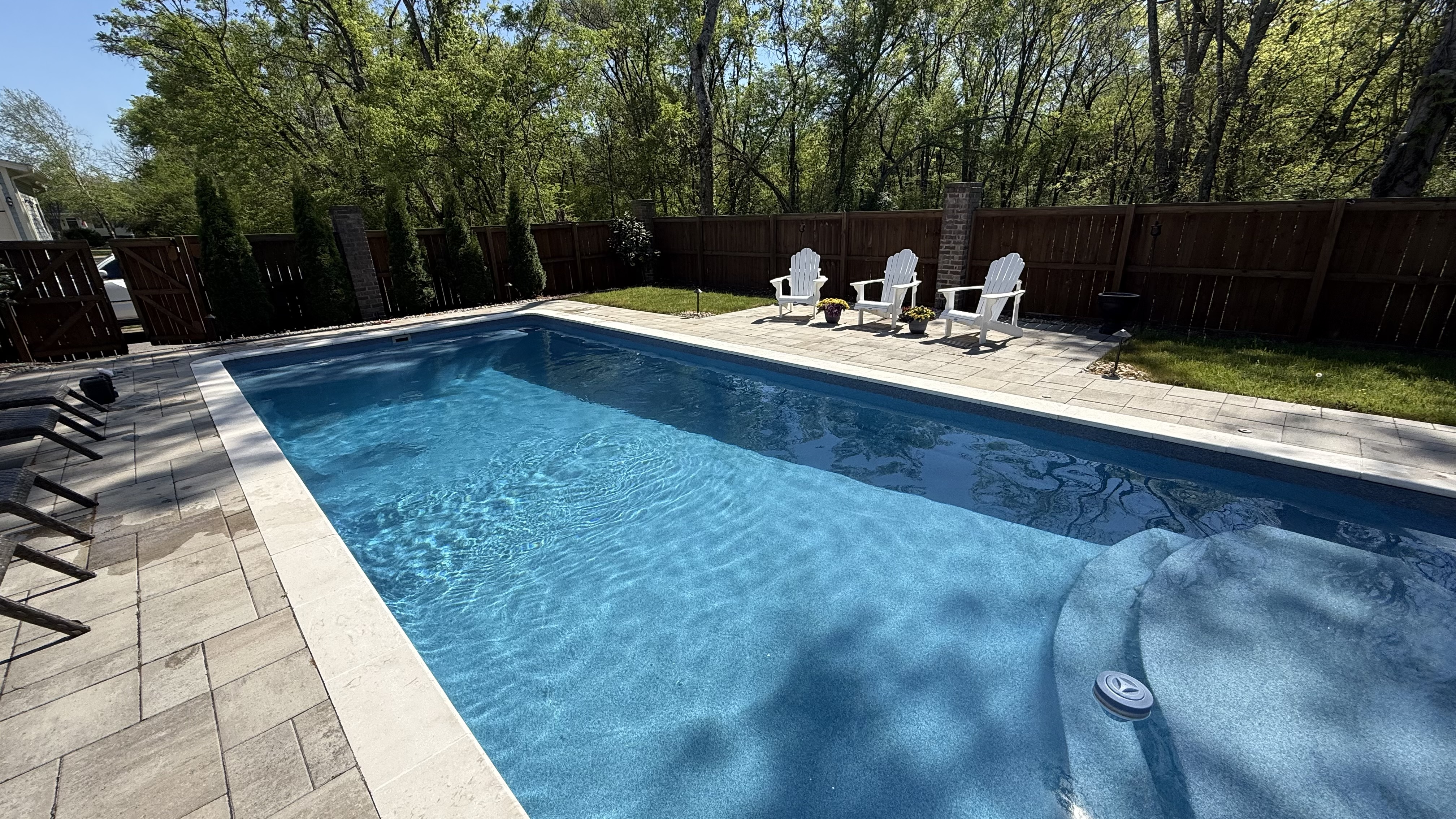Backyard swimming pool with blue water, deck chairs, and a wood fence